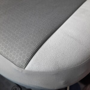 May include: Close-up of a car seat with a two-tone design. The seat features a textured, dark grey fabric on the main seating area, contrasted by a lighter grey, smooth fabric on the side. A grey trim outlines the seat.