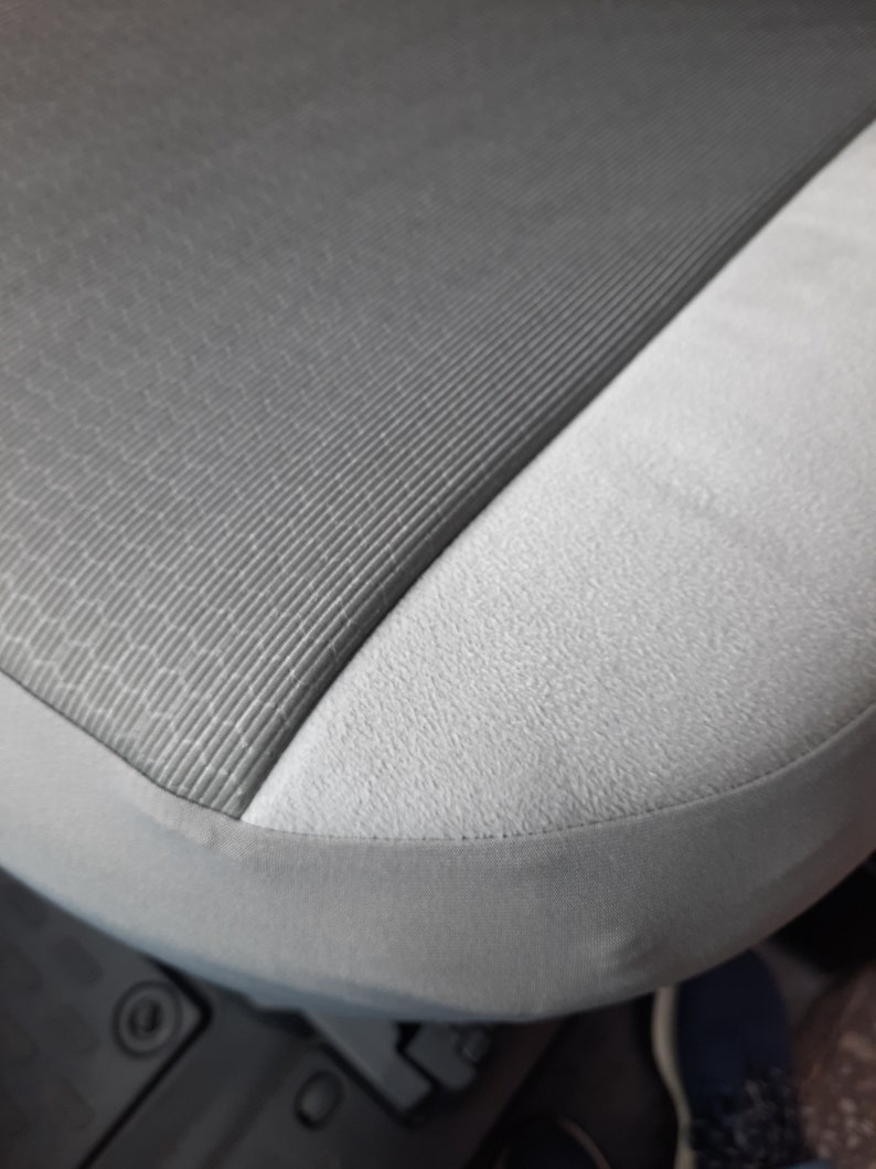 May include: Close-up of a car seat with a two-tone design. The seat features a textured, dark grey fabric on the main seating area, contrasted by a lighter grey, smooth fabric on the side. A grey trim outlines the seat.