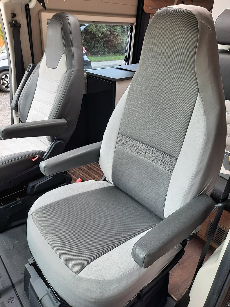 May include: Two-tone grey and white vehicle seats with armrests. The seats feature a textured fabric centre panel and smooth side panels. The armrests are a solid grey colour. The seats are in a vehicle interior.
