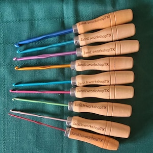 May include: Set of ten crochet hooks with wooden handles. The hooks are in various colors, including blue, purple, orange, green, and red. Each handle is engraved with the text "JandJworkshopTX".