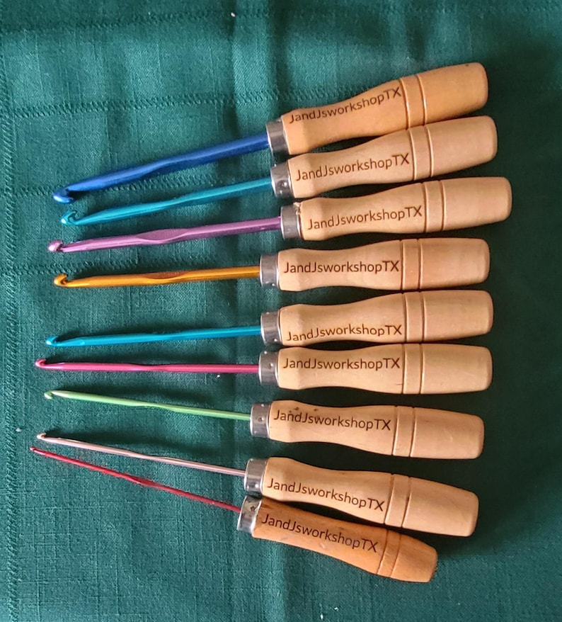 May include: Set of ten crochet hooks with wooden handles. The hooks are in various colors, including blue, purple, orange, green, and red. Each handle is engraved with the text "JandJworkshopTX".