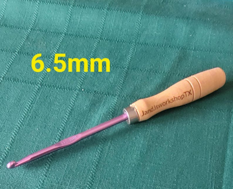 May include: A 6.5 mm crochet hook with a purple metal hook and a wooden handle. The handle is engraved with "JandJworkshopTX".