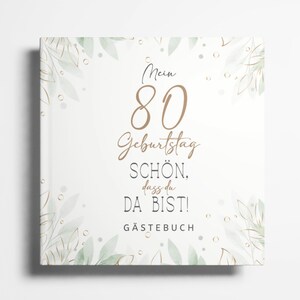 Puede incluir: Un libro de visitas blanco con un diseño floral verde y dorado. El texto de la portada dice "Mein 80 Geburtstag Schön. dass du DA BIST! GÄSTEBUCH".