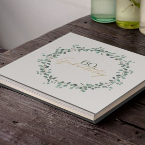 Puede incluir: Un &aacute;lbum de fotos blanco con un dise&ntilde;o de corona de eucalipto verde y el texto "60 Geburtstag" en letras doradas.