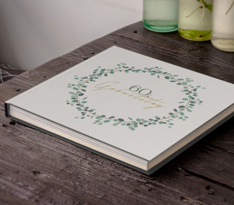 Puede incluir: Un &aacute;lbum de fotos blanco con un dise&ntilde;o de corona de eucalipto verde y el texto "60 Geburtstag" en letras doradas.