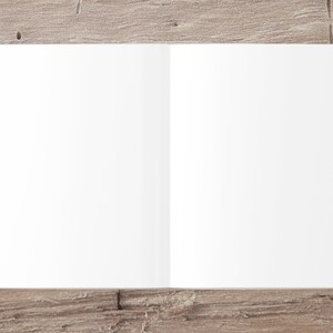 May include: A blank, white, open book lying flat on a light brown wooden surface.