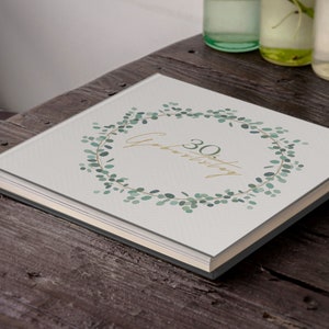 May include: A white photo album with a green eucalyptus wreath design. The text "30th Anniversary" is written in gold lettering inside the wreath.