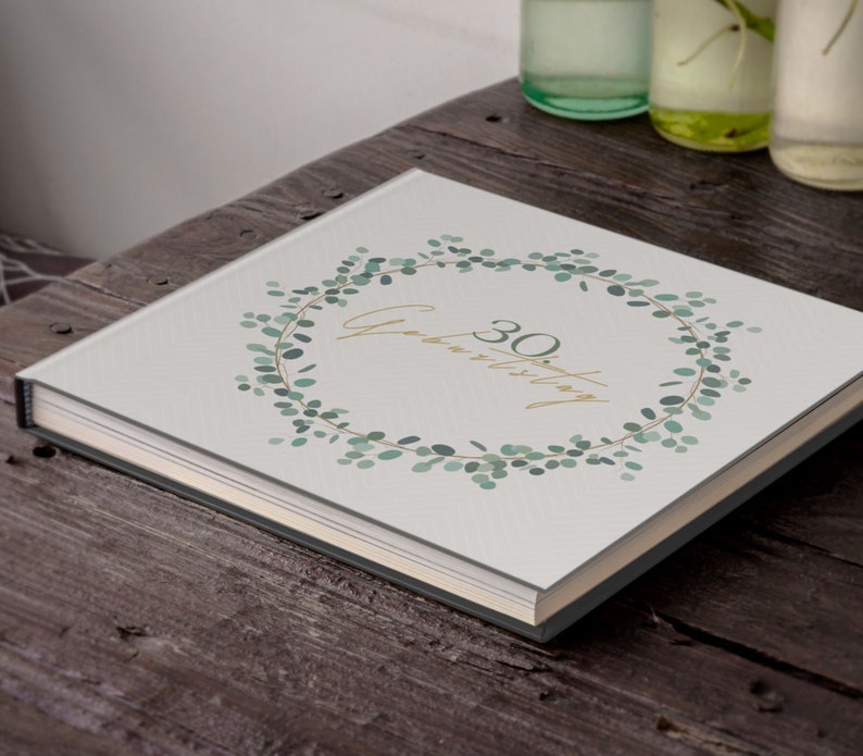 May include: A white photo album with a green eucalyptus wreath design. The text "30th Anniversary" is written in gold lettering inside the wreath.