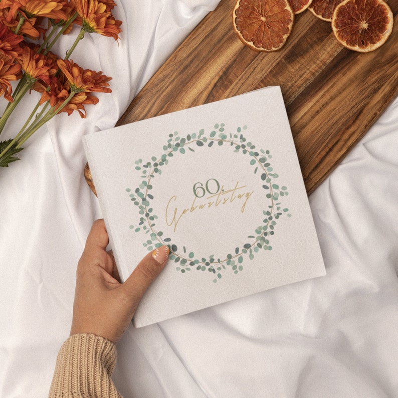 Puede incluir: Una tarjeta blanca con un dise&ntilde;o de corona de hojas verdes y el texto "60. Geburtstag" en letras doradas.