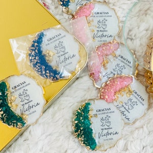 Könnte beinhalten: Vier runde, weiße Untersetzer aus Harz mit goldenen Flocken und einem gewellten Rand. Jeder Untersetzer hat einen anderen Hintergrund: blau, rosa, grün und weiß. Die Untersetzer zeigen ein goldenes Kreuz und eine Tasse mit einer Taube darauf. Der Text auf den Untersetzern lautet "Gracias por acompañarme Mi Bautizo Victoria 11/03/2024".