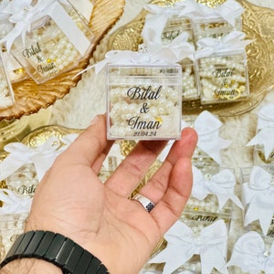 May include: A clear plastic box filled with white pearl beads and a white ribbon bow. The box has a black label with the text "Bilal & Iman 21.04.24".