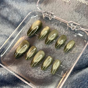 May include: A set of olive green press-on nails in a clear plastic case. The nails have a glossy finish and an oval shape. The case has the text "Bunnies on nails" printed on it.