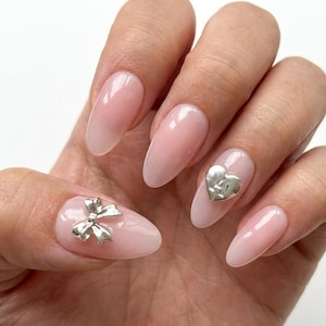 May include: A hand with long, oval-shaped nails. The nails are painted with a light pink color and have silver embellishments, including a bow and a heart. The nails have a glossy finish.