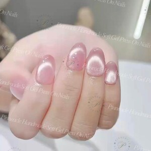 May include: A hand displaying soft gel press-on nails. The nails are oval-shaped and painted in a light pink color with a subtle cat-eye effect. The image includes text that reads "Pinnacle Handmade Soft Gel Press On Nails."