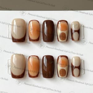 Puede incluir: Un juego de diez uñas postizas con varios diseños y colores. Las uñas presentan una mezcla de tonos marrones, beige y naranjas sólidos, con algunos que muestran patrones abstractos. Las uñas se muestran en una lámina de plástico transparente.