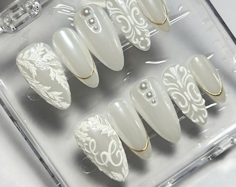 May include: A set of almond-shaped press-on nails in a pearlescent white finish. The nails feature raised white floral and scrollwork patterns, small white pearl accents, and thin gold metallic lines, all arranged within a clear plastic case.