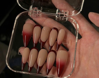 May include: A set of ten almond-shaped press-on nails in a clear plastic storage case. The nails feature a nude base colour with deep red French tips that transition into a gradient ombre effect toward the cuticle area. The case lid has text.