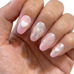 Puede incluir: Una mano con uñas manicuradas. Las uñas son de forma ovalada y están pintadas de color rosa claro con varios diseños. Algunas uñas presentan acentos de corazón blanco, mientras que otras tienen un lazo o un contorno de corazón. La estética general es delicada y femenina.