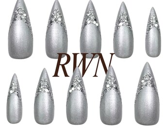 May include: A set of ten stiletto-shaped press-on nails in a metallic silver finish. Each nail features a V-shaped accent of silver glitter at the tip. The letters RWN are printed in a dark brown font in the centre of the arrangement.