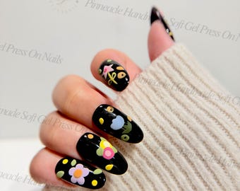 May include: Black oval-shaped soft gel press on nails featuring floral designs in pink, yellow, and blue. The nails have a glossy finish. The text "Pinneade Handmade Soft Gel Press On Nails" is visible.