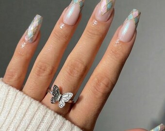 May include: Long coffin-shaped nails featuring a pearlescent nude base with a pastel pink and blue diamond argyle pattern on the tips, outlined in gold glitter. A silver-toned metal ring shaped like a butterfly is worn on the ring finger.