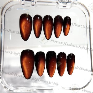 Peut inclure: Un ensemble de dix faux ongles en forme d'amande avec une finition magnétique œil-de-chat brun profond et cuivre. Les ongles sont disposés en deux rangées de cinq dans un boîtier de rangement en plastique transparent portant le texte Pinnacle Handmade Soft Gel Press On Nails.