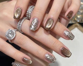 May include: Manicured nails featuring a combination of glossy brown cat-eye polish and silver glitter accents. The fingers are adorned with multiple silver rings, including a prominent round-cut solitaire ring with a halo setting and pavé bands.