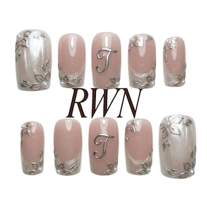 May include: Set of ten artificial nails in various designs. Some nails are pearlescent with silver floral accents. Others are light pink with silver leaf designs and the letter "T". The text "RWN" is at the bottom.