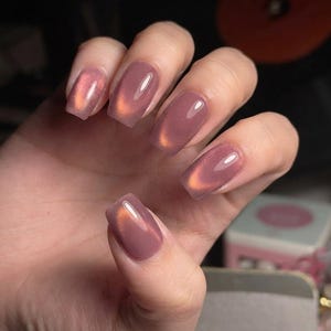 May include: Square-shaped fingernails featuring a glossy, mauve-toned cat-eye polish. The finish displays a shimmering, metallic copper-colored streak that runs diagonally across the center of each nail, creating a reflective, dimensional effect.