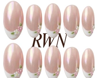 May include: A set of ten oval-shaped press-on nails with a pearlescent light pink base and white French tips. Each nail features small, hand-painted white and pink flowers with green leaves. The text RWN and Rich Wife's Nails by Pinneacle appear.