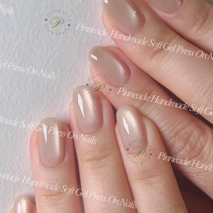 May include: Close-up of a hand with almond-shaped, light beige press-on nails. The nails have a glossy finish and a subtle shimmer. The text "Pinneacle Handmade Soft Gel Press On Nails" is visible in the background.