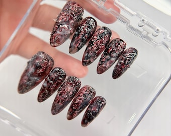 May include: A set of ten almond-shaped press-on nails featuring a textured, multi-dimensional finish in deep shades of crimson red, black, and white. The surface has a rugged, cratered appearance with a glossy top coat, arranged in a clear plastic case.