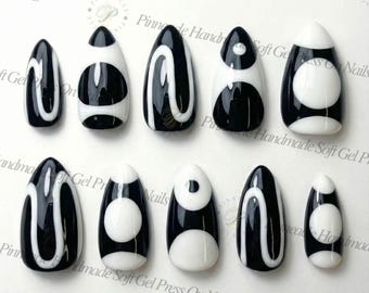May include: A collection of ten black and white press-on nails. Each nail has a glossy finish with diverse designs, including swirls, circles, and geometric patterns. The nails are almond-shaped and displayed on a white surface. The text "Pinnacle Handmade Soft Gel Press On Nails" is visible.