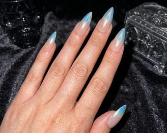 May include: A hand with long, pointed almond-shaped nails featuring a clear base and a shimmering, iridescent blue holographic tip. The nails have a smooth, glossy finish that reflects light, creating a subtle colour-shifting effect on the tips.
