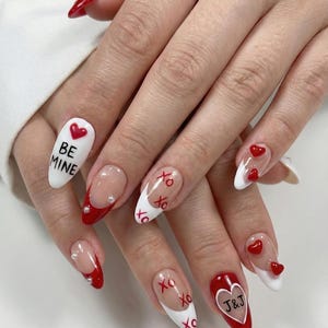 May include: A set of Valentine's Day-themed nail art. The nails feature a variety of designs, including red tips, white bases with red hearts, the words "BE MINE", and "XO" lettering. Some nails have a clear base with red heart accents and the initials "J&J".