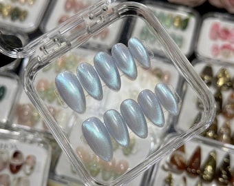 May include: A set of ten almond-shaped press-on nails with a pearlescent, light blue cat-eye finish. The nails are arranged in two rows of five inside a clear, square plastic storage case, showcasing a reflective, shimmering surface texture.
