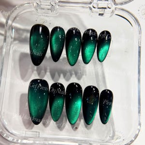 May include: Set of emerald green press-on nails with a cat-eye effect. The nails have a glossy finish and a gradient design, transitioning from black to green. The nails are in a clear plastic case.
