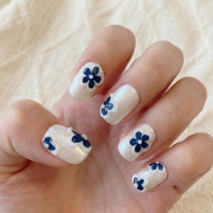 May include: White nail polish with dark blue floral designs. Each nail features a different arrangement of the flowers, creating a cohesive yet unique look. The nails are oval-shaped and have a glossy finish.