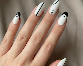 May include: Oval-shaped nails featuring a white base coat with black accents. The design includes black tips, thin vertical lines, and small black dots. The nail art is applied to five fingers, showcasing a clean, high-contrast aesthetic with precise lines.