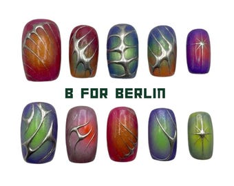 May include: A set of ten press-on nails featuring gradient colours in shades of pink, orange, blue, purple, and green. The nails are decorated with raised silver metallic liquid-style patterns and starburst designs. Text reads B FOR BERLIN BY PINNEACLE.