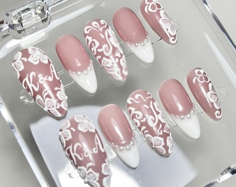 May include: A set of ten almond-shaped press-on nails featuring a dusty rose base colour. Some nails display white floral patterns and the letters K&A, while others feature a white French tip accented with a row of small white pearls at the base.
