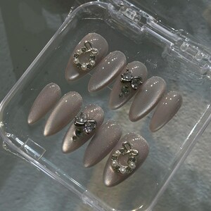 May include: A set of artificial nails in a clear plastic case. The nails are a light, shimmery taupe color. Some nails feature silver bow accents with clear rhinestones, while others have silver wreath accents with small white pearls.
