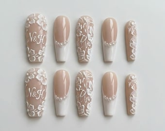 May include: A set of ten press-on nails featuring a nude base with white 3D floral relief designs, white French tips accented with small white pearls, and the letters N&H embossed in white on the thumb nails, arranged in two rows of five.