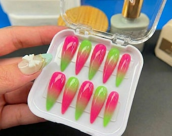 May include: A set of ten stiletto-shaped press-on nails featuring a vibrant gradient design that transitions from bright pink at the tip to lime green at the base. The nails are arranged in a white plastic storage container with a clear hinged lid.