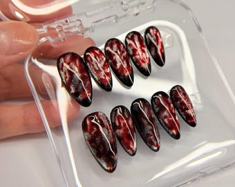 May include: A set of ten almond-shaped press-on nails featuring a deep red and black gradient base with a shimmering, magnetic cat-eye effect and thin, white, vein-like marbled lines running across the surface, displayed in a clear plastic case.