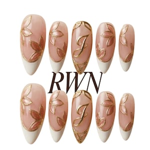 Puede incluir: Un conjunto de diez uñas artificiales con base rosa claro, puntas blancas y diseños florales e iniciales dorados. Las uñas tienen forma de almendra y presentan las letras "RWN" en fuente marrón oscuro.