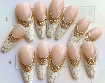 May include: A set of ten press-on nails featuring a nude base, a white crackle pattern with gold veining, and a gold glitter crescent accent at the cuticle. The nails are arranged in two rows labelled L and R, showcasing a long, tapered coffin shape.
