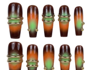 May include: A set of ten press-on nails featuring a gradient design that transitions from dark brown at the tips to a bright green centre and orange base. The nails are adorned with metallic gold rings and small gold spheres attached to the surface.