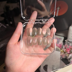 May include: A set of ten clear, iridescent press-on nails with a coffin shape, arranged in two rows of five inside a small, square, transparent plastic storage case with a hinged lid and a secure latch closure mechanism.