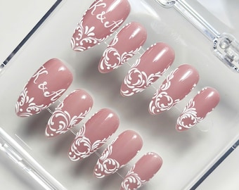 May include: A set of ten almond-shaped press-on nails in a dusty rose pink colour. Each nail features a glossy finish with intricate white filigree scrollwork patterns. Two of the nails are prominently marked with the white text K & A.